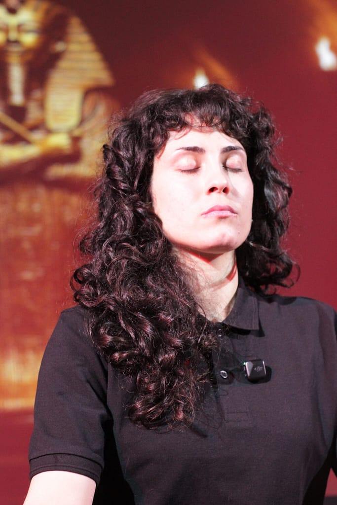 actriz teatro ingles escena inmersiva secundaria scaled Mujer con pelo rizado y ojos cerrados, con micrófono, en una escena de teatro en inglés con fondo egipcio.
