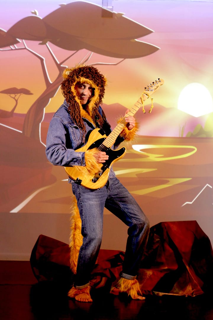 Dora Do-Much: Obra de Teatro Musical con Animales en Inglés León rockero tocando la guitarra eléctrica. Escenario de sabana africana para teatro infantil ODS.